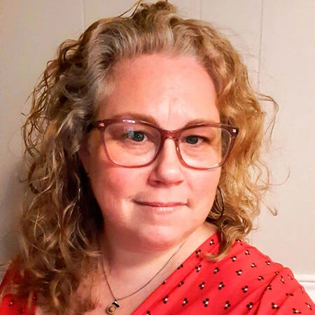 Auteure Martine Ouellet | DistributionDPA Woman with curly blonde hair wearing red-framed glasses and a red patterned blouse, facing the camera with a slight smile.